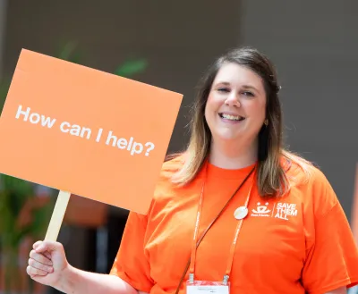 Smiling volunteer holding a sign that says "How can I help?"