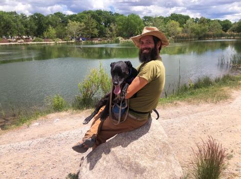 Ken wearing a cowboy hat and holding Minnie the dog in his lap, sitting on a boulder in front of a river