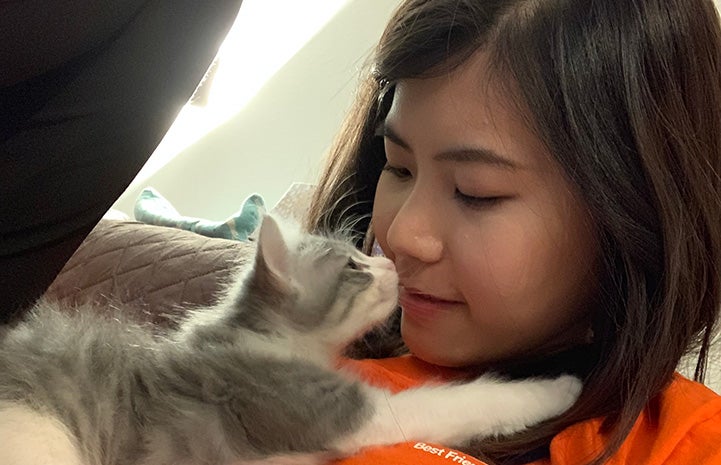 Woman lying down with a gray and white tabby kitten stretched out on her sniffing her nose