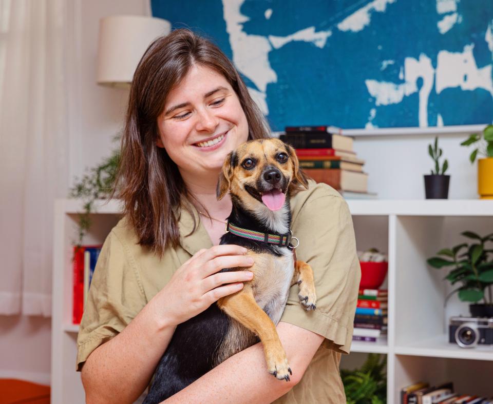 Smiling person holding a small dog in front of a book case with a painting above it